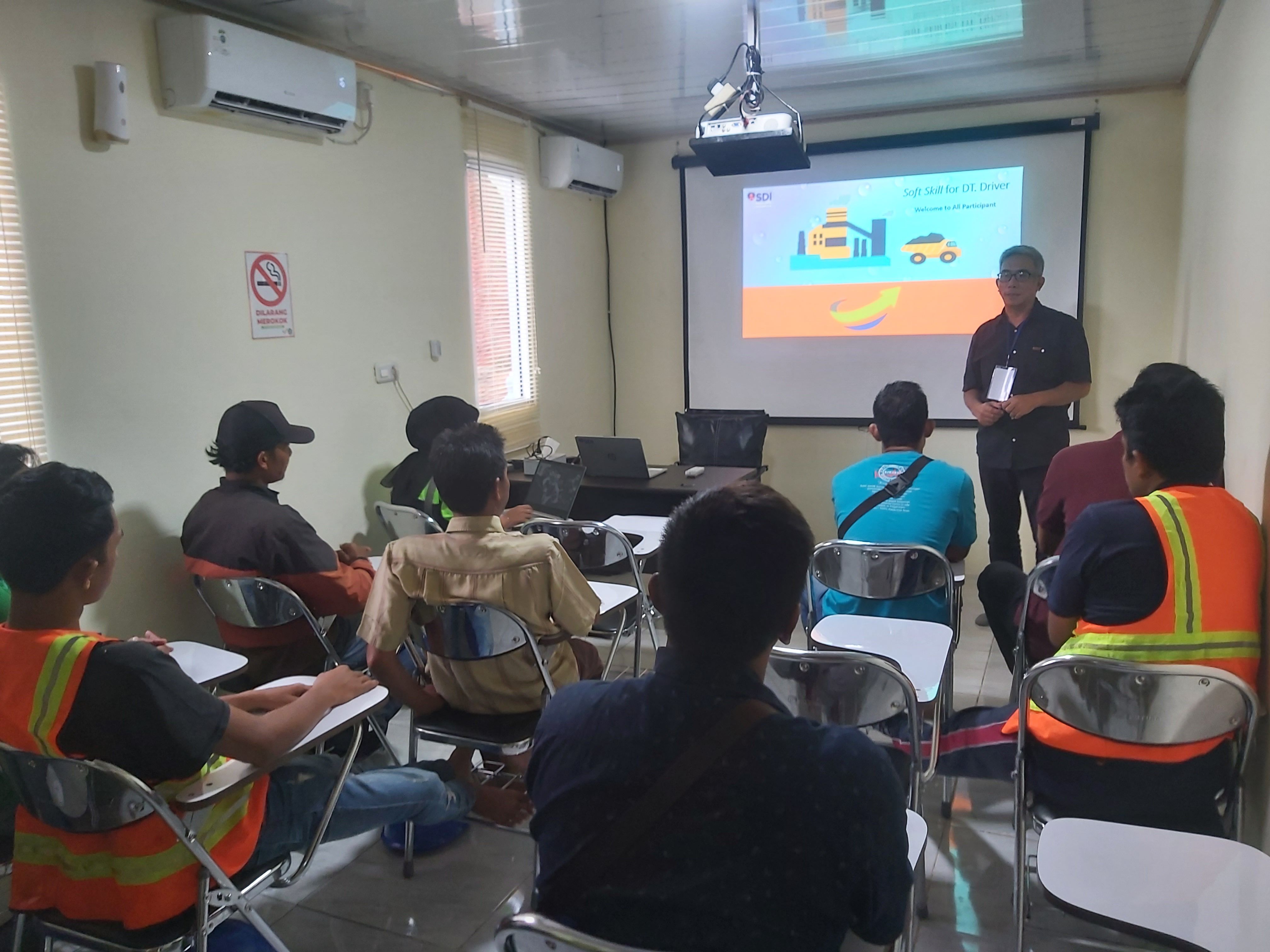 Pelatih SDI berdiskusi dengan peserta berseragam kerja dan rompi keselamatan di ruang pelatihan